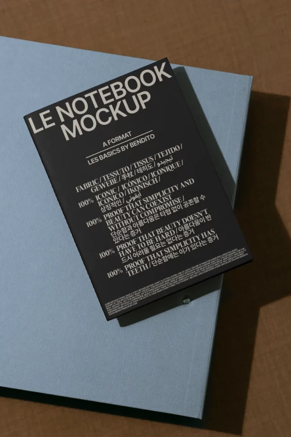 A black notebook titled LE NOTEBOOK MOCKUP rests at an angle on a closed blue book, displayed on a brown surface with angled lighting casting subtle shadows.