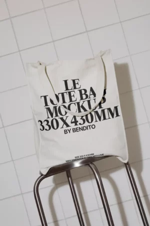 A white tote bag with black text reading LE TOTE BAG MOCKUP 330X430MM BY BENDITO rests on the back of a chair, set against a tiled wall.