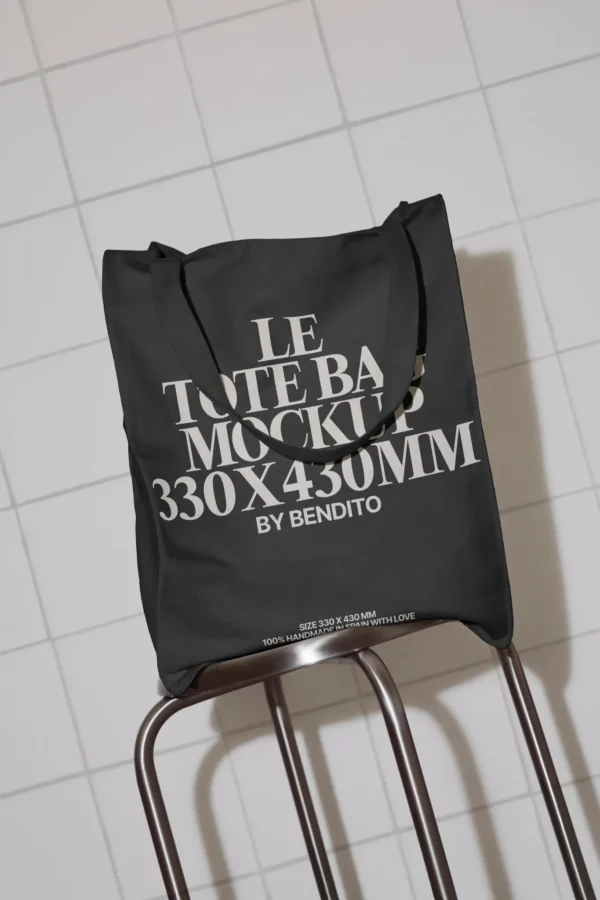 A black tote bag with bold white text reading LE TOTE BAG MOCKUP 330x430MM BY BENDITO hangs on the back of a metal chair against a white tiled wall.