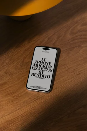 A smartphone with a mockup design displayed on the screen lies on a wooden surface, illuminated by natural light from the left. The screen shows bold text in French about the iPhone mockup details.