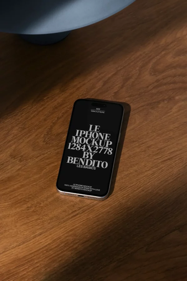 An iPhone lies face-up on a wooden surface in sunlight, displaying a mockup screen with bold white text that reads, Le iPhone Mockup 1284 x 2778 by Bendito Les Basics.