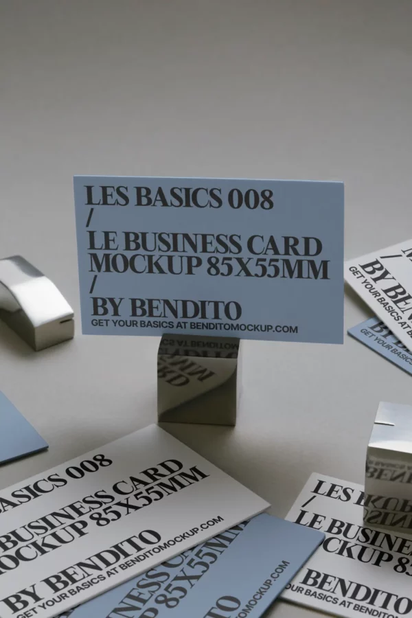 A blue business card reading LES BASICS 008 / LE BUSINESS CARD MOCKUP 85X55MM // BY BENDITO is clipped upright, surrounded by similar cards and metallic cardholders on a gray surface.