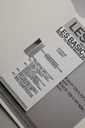 A close-up of folded white and gray printed packaging with black text, displaying “LES BASICS READY TO WEAR MOCKUPS” and product information, placed on a neutral surface with overlapping paper sheets.