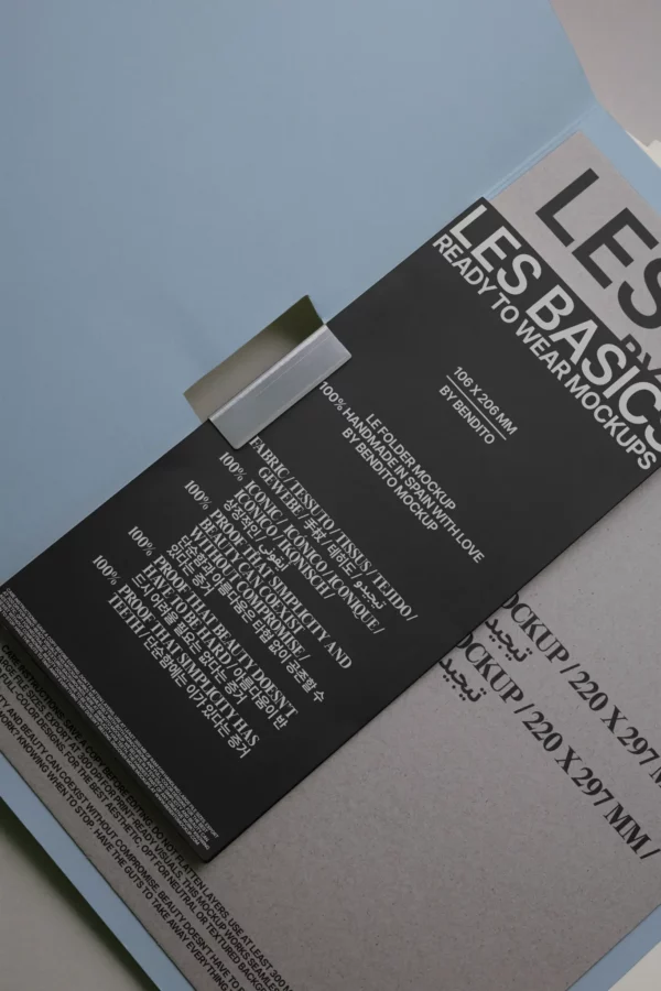 A black clothing tag with white text displaying material information and washing instructions is attached to a light blue and beige folder or package. The tag reads LES BASICS READY TO WEAR MOCKUPS.