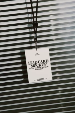 A white ID card mockup with a black lanyard hangs in front of horizontal blinds. The card displays bold black text reading “LE ID CARD MOCKUP” and other details, all in a modern, minimalist design.
