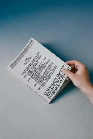 A hand holds a magazine with a white cover featuring bold black text in various fonts and languages on a gray and blue gradient background. The magazine is tilted diagonally on a flat surface.