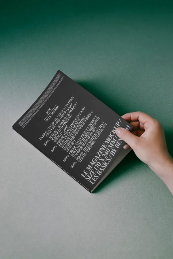 A hand holds a black book with large white text on the cover, placed on a green and gray gradient surface. The text is arranged in a scattered, overlapping layout.