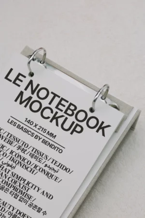A ring-bound notebook mockup with a cover page titled LE NOTEBOOK MOCKUP in bold black letters, featuring various text samples in different fonts and languages on a light, textured background.