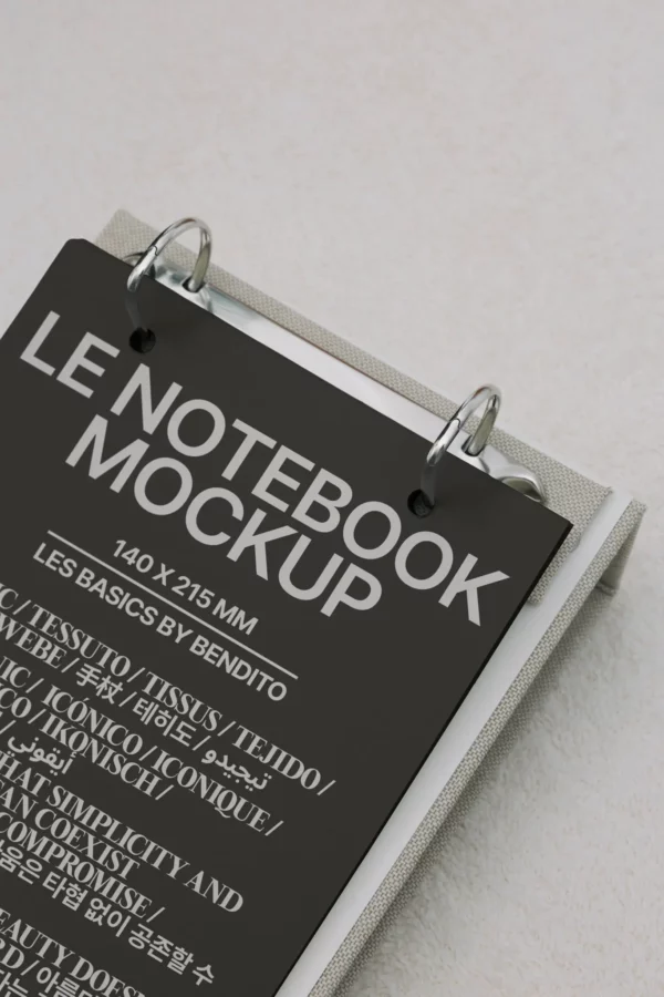 A closeup of a ring-bound notebook with a black cover displaying the text LE NOTEBOOK MOCKUP 140 × 215 MM in bold white letters and additional multilingual text beneath, resting on a light surface.