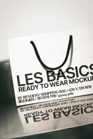 A white shopping bag with black rope handles displays bold black text reading LES BASICS READY TO WEAR MOCKUP along with size details and multilingual print, standing on a reflective metallic surface.