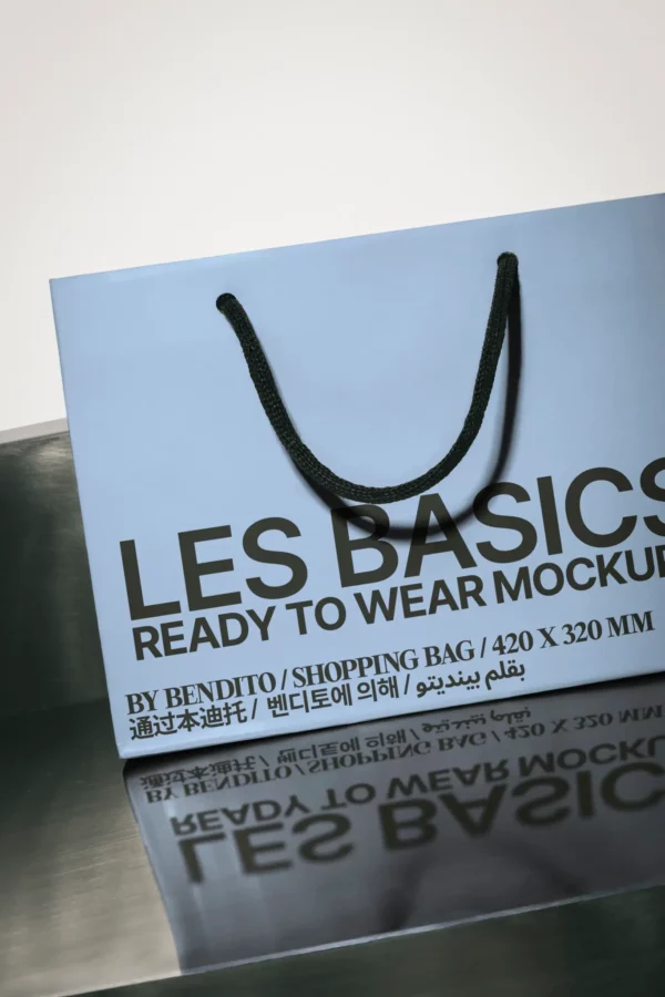 A light blue shopping bag with black rope handles sits on a reflective surface. Bold black text on the bag reads LES BASICS READY TO WEAR MOCKUP along with product details in multiple languages.