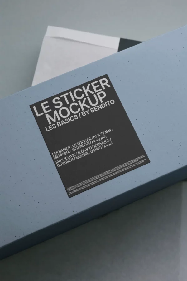A blue rectangular box with a black and white label on top displaying the text LE STICKER MOCKUP LES BASICS | BY BENDITO along with smaller detailed information beneath it, photographed on a gray surface.