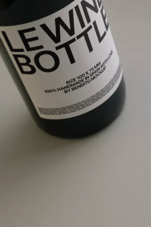 A close-up of a wine bottle with a white label that reads LE WINE BOTTLE in large black letters. Additional text below notes the size and that it is handmade in Spain by Bendito Mockup.