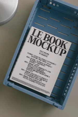 A white book mockup with bold black text titled LE BOOK MOCKUP rests inside a blue plastic basket on a light gray surface. The book lists fabric types and features in both English and Korean.