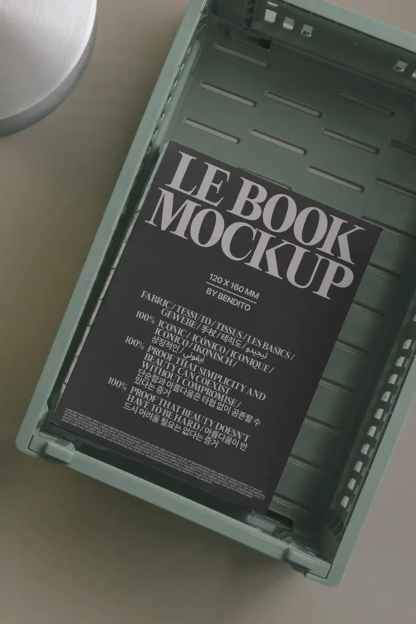 A black book mockup with white text labeled LE BOOK MOCKUP rests inside a green plastic crate with slotted sides. Part of a round metallic object is visible on the left.