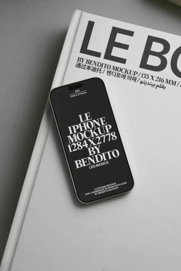 A black iPhone displaying mockup text rests on a large white hardcover book with bold black and gray text partially visible on the cover.
