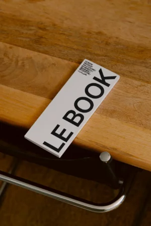 A booklet titled LE BOOK with bold black letters lies on a wooden table next to a black chair.