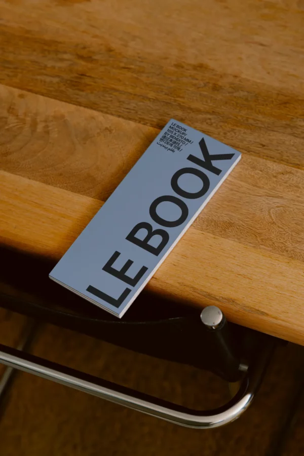 A booklet titled LE BOOK with large black text lies on a wooden table next to a black chair with a metal frame.