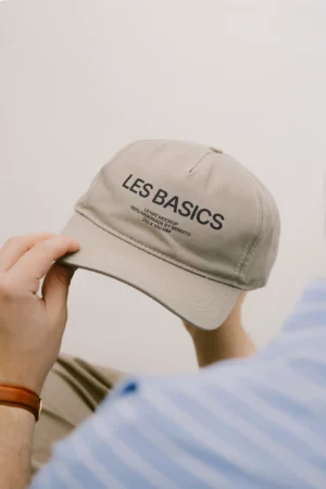 A person wearing a striped blue shirt and a brown watch holds a beige baseball cap with the words LES BASICS printed in black letters on the front. The background is plain and light-colored.