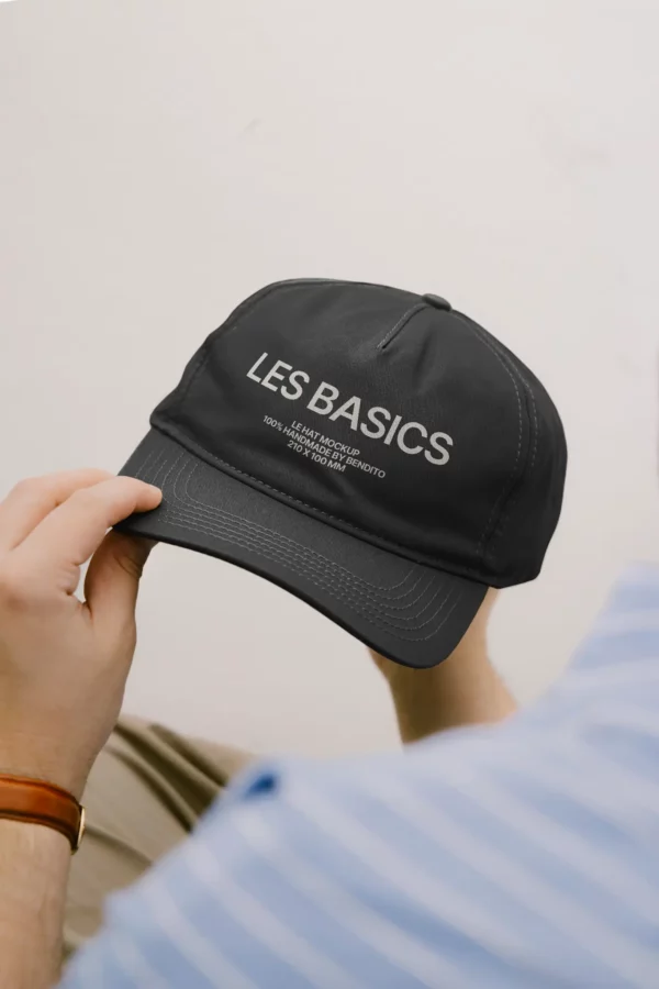 A person holding a black baseball cap with LES BASICS and smaller white text printed on the front. The person wears a blue and white striped shirt and a brown watch. The background is a plain light color.