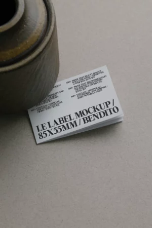 A stack of white business cards with bold black text reading LE LABEL MOCKUP / 85X55MM / BENDITO, partially covered by a round wooden object on a light grey surface.