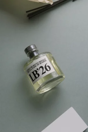 A clear glass perfume bottle labeled LB 26 with a metallic cap is lying on a light green surface. The bottle is partially filled with a pale yellow liquid.