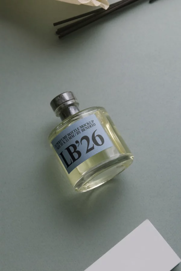 A clear glass bottle filled with a pale yellow liquid labeled LB26 is lying on its side on a gray-green surface, with part of a white paper and dark sticks visible nearby.