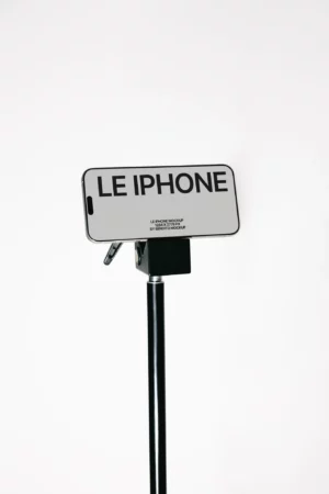 A smartphone on a tripod displays a screen with large text reading LE IPHONE against a plain white background.