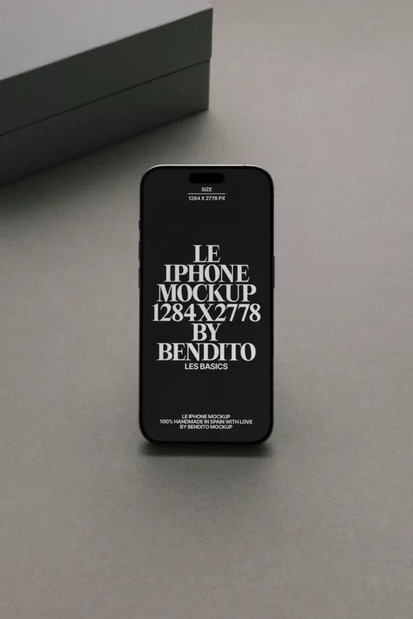 A black smartphone displaying white text, standing upright on a gray surface with a gray box in the background. The screen text reads “LE IPHONE MOCKUP 1284x2778 BY BENDITO LES BASICS” in bold letters.