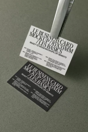 Two business cards, one white and one black, are displayed on a grey surface. The cards feature bold, uppercase text with mockup details, and the white card is held by a metal clip above the black card.