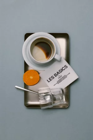 A cup of coffee, a half orange, a glass with two spoons, and a booklet labeled LES BASICS are arranged neatly on a rectangular metal tray against a gray background.