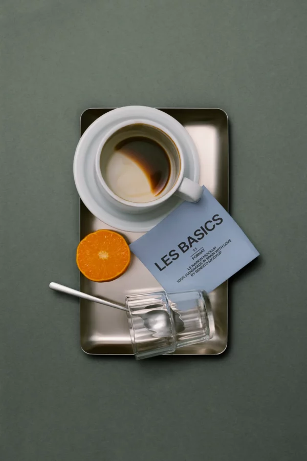 A white coffee cup with espresso on a metal tray, next to half an orange, a glass cup with a spoon, and a card labeled LES BASICS on a gray background.