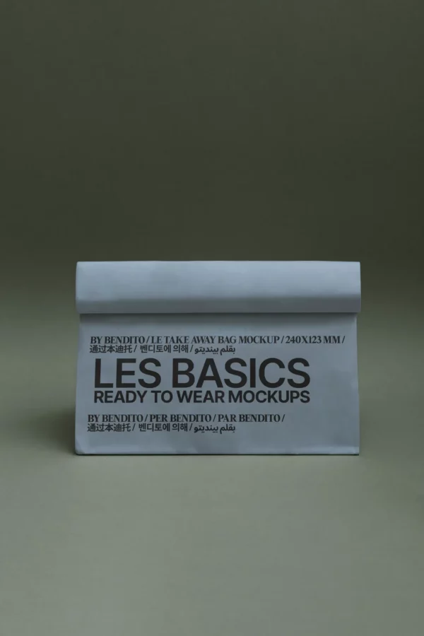 A white, folded paper bag stands upright against a greenish-gray background. The bag displays bold black text that reads LES BASICS READY TO WEAR MOCKUPS along with additional details in multiple languages.