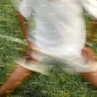 A person in a white shirt, white shorts, and a white cap is moving quickly on a grassy field, creating a motion blur effect. The person appears to be exercising or warming up.