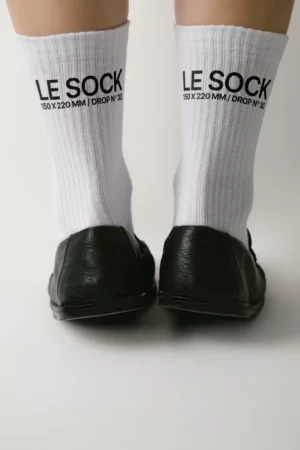 Back view of a person wearing white ribbed socks with black text that reads “LE SOCK 150 x 220 MM / DROP N° 30” and black shoes, standing on a plain light background.