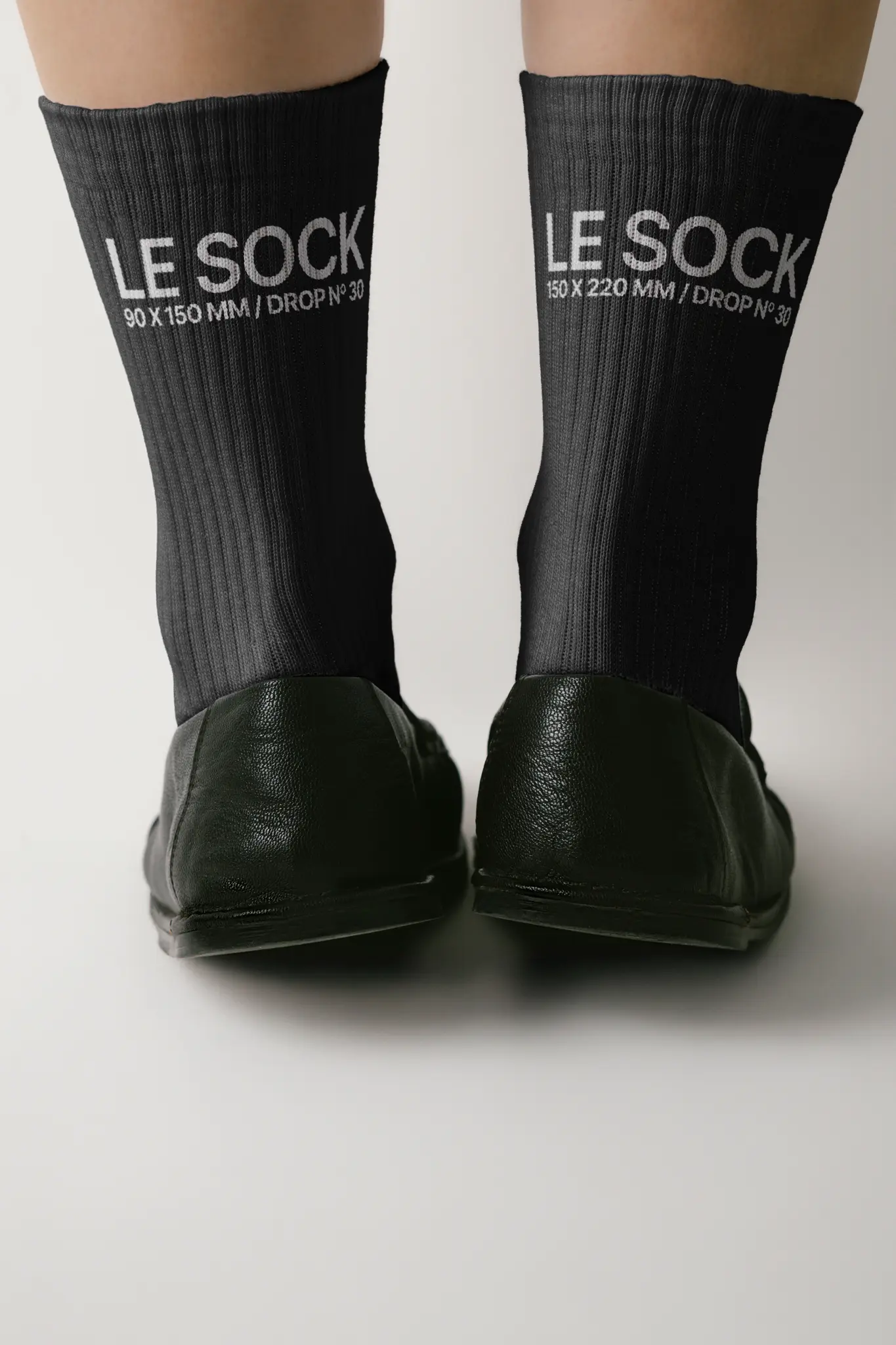 Back view of black socks with white text LE SOCK 90 x 150 MM / DROP Nº 30 and LE SOCK 150 x 220 MM / DROP Nº 30, worn with black shoes, against a plain background.