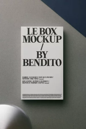 A rectangular white box with bold black text that reads “LE BOX MOCKUP / BY BENDITO” lies on a gray surface next to a dark blue area and a round white object in the corner. Smaller descriptive text appears at the bottom of the box.