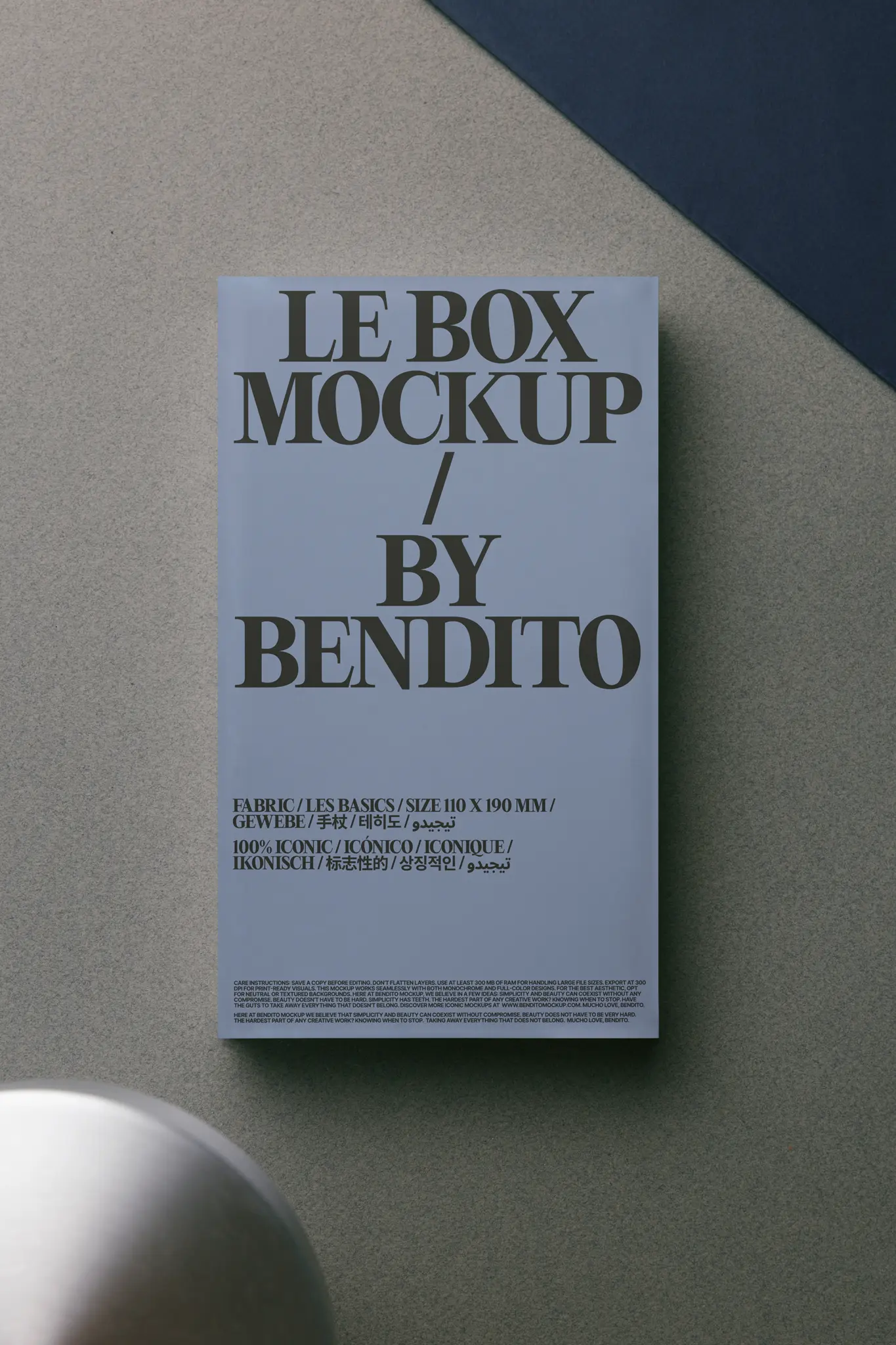 A rectangular poster with large text reading LE BOX MOCKUP / BY BENDITO lies on a textured gray surface, partially overlapping a dark blue area, with a blurred round object in the bottom left corner.