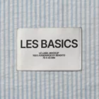 A white cloth label with black text reading LES BASICS LE LABEL MOCKUP 100% HANDMADE BY BENDITO 70 X 45 MM is sewn onto light blue and white vertically striped fabric.