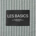 A black rectangular clothing label with the text LES BASICS stitched onto a green and white vertical striped fabric background. Additional smaller text reads: LE LABEL MOCKUP 100% HANDMADE BY BENDITO 70 X 45 MM.