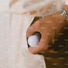 A close-up of a person’s hand holding a golf ball, wearing a silver watch and dressed in light-colored clothing. The image has a watermark reading BENDITO repeated across it.