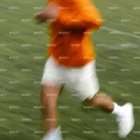 A blurred image of a person in an orange hoodie, white shorts, and white shoes running on a grassy field. The image captures a sense of motion and speed.