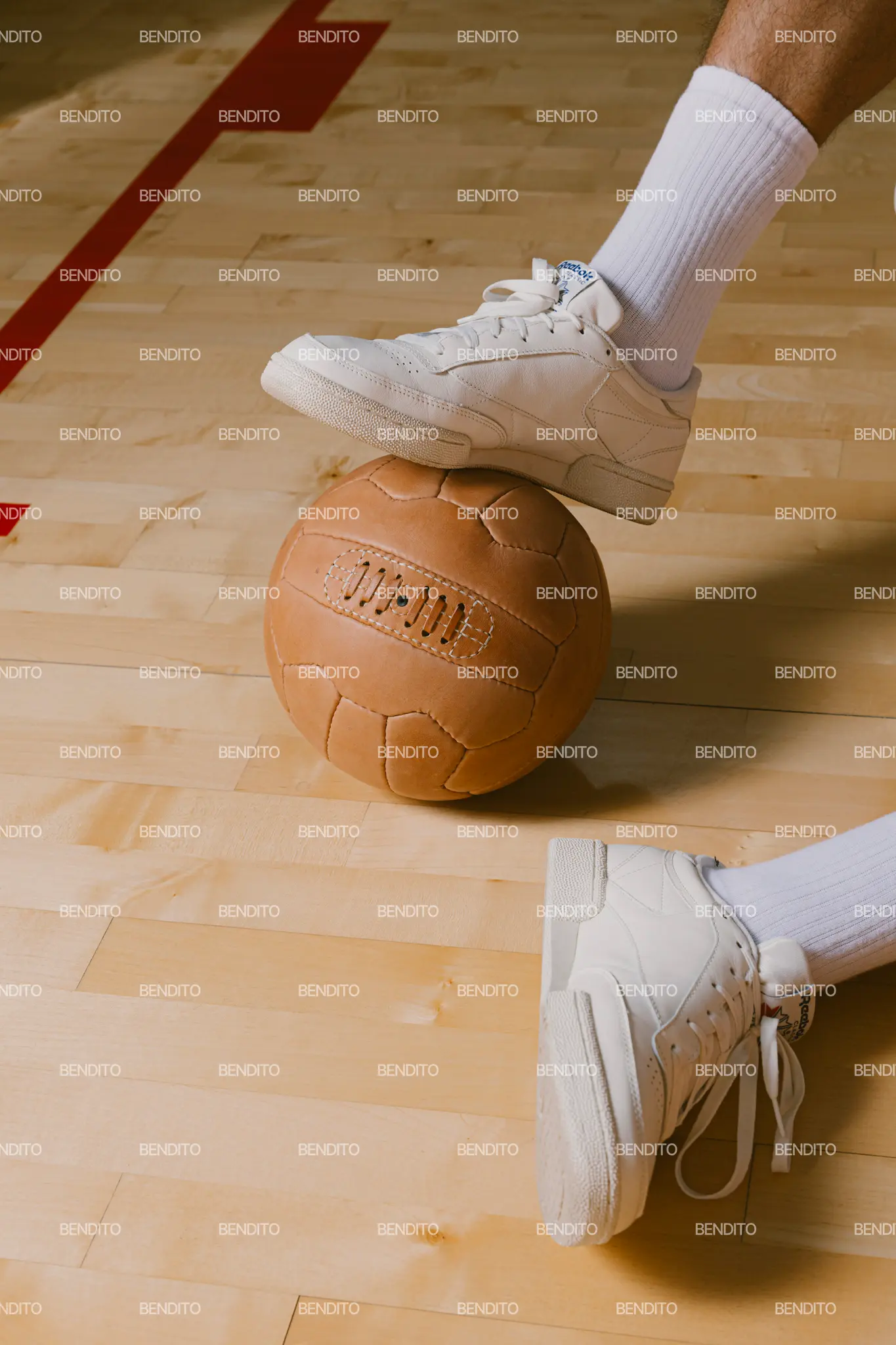 A person wearing white sneakers and white socks balances one foot on top of a brown basketball on a wooden indoor court, with a red line visible in the background.