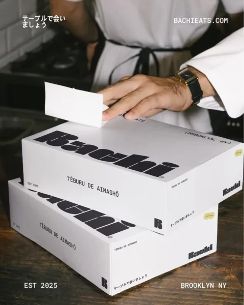 A person wearing an apron seals a stack of three white food boxes labeled Bachi and TĒBURU DE AIMASHŌ. The boxes are on a table, with text reading EST 2025 and BROOKLYN NY.
