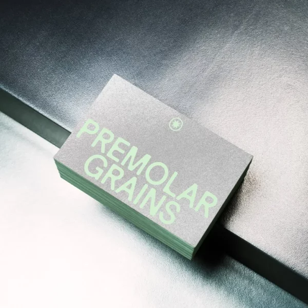 A stack of gray business cards with large, light green text reading PREMOLAR GRAINS sits on a metallic surface under bright lighting.