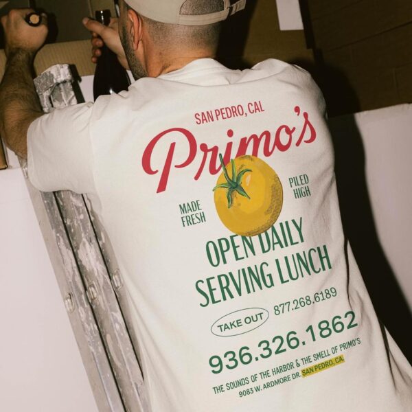 A man in a cap stands on a ladder with his back to the camera, wearing a white Primo’s t-shirt advertising a San Pedro, CA restaurant, featuring a tomato graphic and contact information.