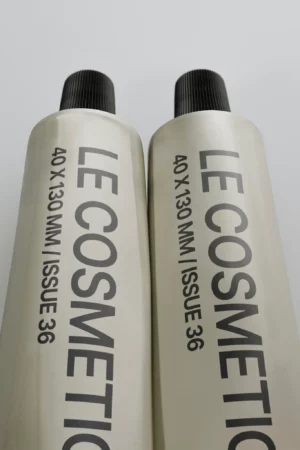 Two metallic tubes with black caps are placed side by side on a light surface. The tubes are labeled LE COSMETIC 40 X 130 MM / ISSUE 36 in bold, black letters running lengthwise along each tube.