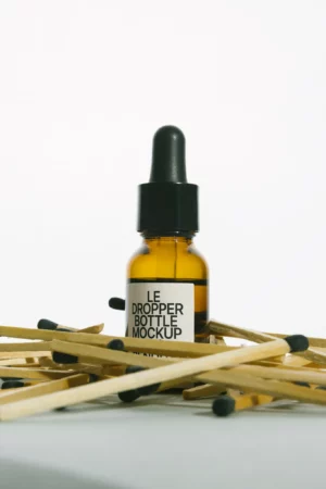 A small amber dropper bottle labeled LE DROPPER BOTTLE MOCKUP stands upright among scattered wooden matches with black tips on a light surface, against a plain white background.