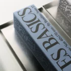 A rectangular blue box with large black text reading “BASICS” and smaller text covering its sides sits on a reflective metal tray, with part of another similar box visible in the background.