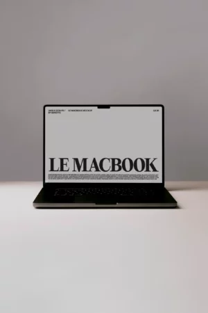 A black laptop sits on a white surface against a gray background, displaying a screen with large text that reads LE MACBOOK in bold letters.